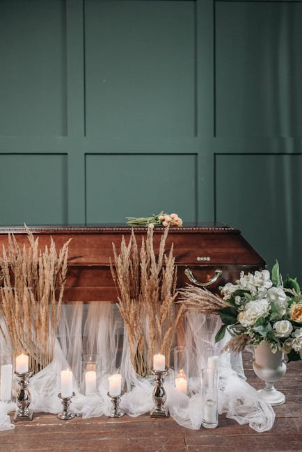 A floral arrangement featuring white roses, peonies, and layered green leaves in a white ceramic vase, positioned on a wooden floor next to a vintage wooden piano. Surrounding the vase are tall, beige