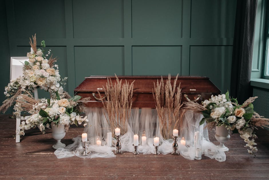 A funeral floral arrangement display set against a dark green paneled wall, featuring two large white vases filled with cream and white roses, hydrangeas, and soft peach blooms, complemented by tall,