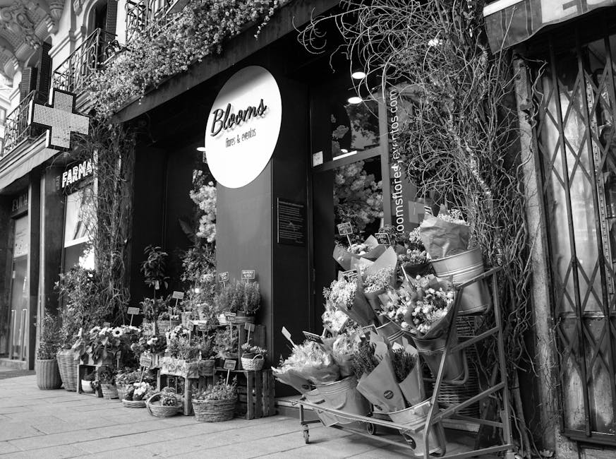 A flower shop front with a large illuminated circular sign reading 'Blooms Flowers & Essentials', surrounded by an array of floral arrangements and potted plants displayed on wooden crates and metal c